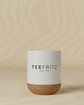 Weiße Teetasse mit Korkboden, darauf steht "TEEFRITZ BIO-TEE", vor einem beigen Hintergrund.